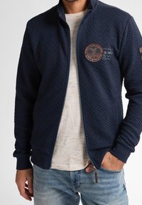 Marinblå kviltad zip-up jacka med strukturerat mönster, som har en broderad logotyp och kontrasterande ljusblå detaljer. Bärs över en ljus skjorta.