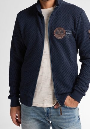 Chaqueta Navy acolchada con cremallera y patrón texturizado, que presenta un logo bordado y un ribete en azul claro. Lleva puesta sobre una camisa clara.