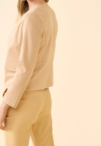Blazer beige cintré à manches longues, col plat et poches latérales, associé à un pantalon assorti jaune clair, tissu à texture lisse.