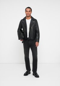 Chaqueta de motorista de cuero negro con cremalleras plateadas, combinada con una camiseta blanca y jeans oscuros. Modelo sosteniendo un bolso negro, de pie sobre un fondo blanco.