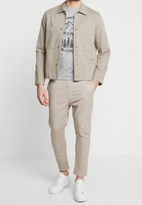 Beige jas met twee borstzakken, grijs T-shirt met bergafbeelding, en taps toelopende beige broek. Afgemaakt met witte sneakers.