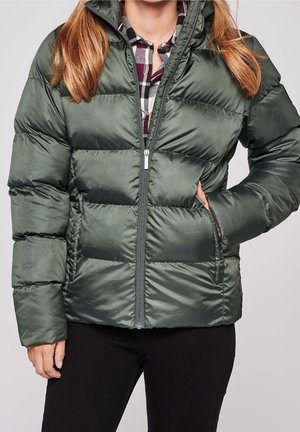 Femme portant une doudoune matelassée vert foncé sur une chemise à carreaux, une main dans la poche de la veste et un pantalon noir.