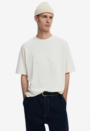 T-shirt in cotone crema con logo in rilievo "DESIGUAL EST. 1984". Abbinato a pantaloni in denim scuro e a un berretto di maglia chiara.