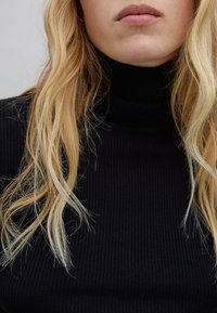 Maglione nero a collo alto con una vestibilità aderente. Tessuto fine e morbido. Lunghi capelli biondi mossi parzialmente visibili.