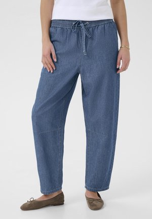 Kvinde iført løstsiddende blå denimbukser med snøre og brune balletsko, stående foran en ensfarvet baggrund.