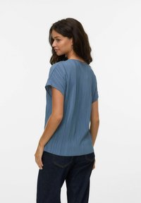 Donna con capelli scuri, che indossa un top blu a maniche corte plissé e jeans scuri, in piedi con la schiena rivolta e la testa leggermente girata a sinistra.