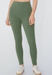 Legginsy z wysokim stanem w kolorze oliwkowym, wykonane z elastycznej tkaniny; gładka tekstura; dopasowany krój z długością do kostek, zestawione z białymi trampkami.