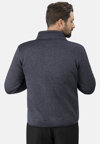 Mann mit kurzem braunem Haar und Bart, der eine strukturierte dunkelblaue Jacke trägt, von hinten gegen einen schlichten hellen Hintergrund gezeigt.