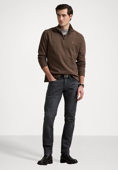 Un pull zippé marron sur une chemise grise, associé à un jean gris foncé et des bottines noires. Le modèle adopte une pose relaxée.