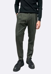 Pantaloni verde oliva con vestibilità affusolata, pieghe frontali e vita con coulisse, abbinati a sneakers nere. Tessuto liscio e design minimalista.