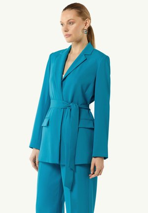 Teal blazer made of smooth fabric with a fitted design, long sleeves, a belted waist, and two front pockets. Complementary teal trousers.