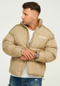 Beige gepolsterte Jacke mit hohem Kragen, Reißverschluss und Logo auf der Brust. Getragen über einem weißen T-Shirt, mit blauen verwaschenen Jeans und einer Uhr.
