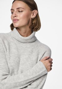 Jauna sieviete ar īsiem brūniem matiem, valkājot zelta auskarus un gaisīgi pelēku ribveida augstu apkaklīti, skatās sānis un ir sakrustotas rokas.