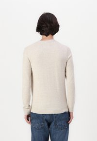 Beige kötött pulóver hosszú ujjakkal és kerek nyakkal. A pulóver bordázott aljjal és mandzsettával rendelkezik. Kék farmernadrág felett viselik.