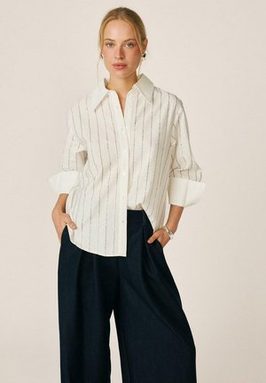 Mujer con cabello rubio, con una camisa blanca suelta abotonada con finas rayas verticales y pantalones anchos oscuros, manos en los bolsillos, de pie frente a un fondo liso.