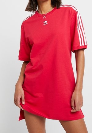 Femme portant une robe T-shirt Adidas rouge avec des bandes blanches sur les manches et le logo Adidas sur la poitrine, tenant l'ourlet de la robe.
