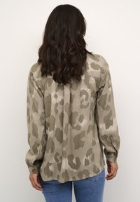 Camisa de manga larga en caqui con un estampado de leopardo. Presenta puños abotonados y una espalda dividida para facilitar el movimiento.
