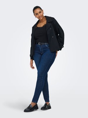 Ženska, oblečena v črno kratko jakno, črno zgornjo majico z čipkasto obrobo, temno modre ozke kavbojke, črni pas in črne mokasine, stoji z prekrižanimi nogami.