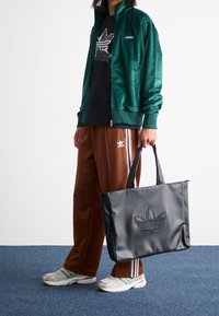 Grüne Velourjacke, schwarzes T-Shirt, braune Trainingshose mit weißen Streifen und eine schwarze strukturierte Tragetasche mit geprägtem Logo.