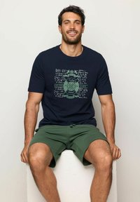 T-shirt blu navy con un design grafico verde contenente testo ed elementi marini. Abbinato a pantaloni corti verdi, modello seduto su un blocco bianco.