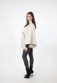 Kremowa peleryna z wysokim kołnierzem i wiązaniem w talii, zestawiona z czarną koszulą z długim rękawem, szarymi obcisłymi dżinsami i czarnymi botkami na wysokim obcasie.