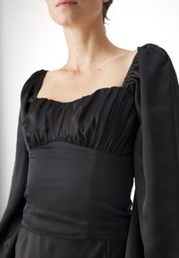 Abito nero con scollatura quadrata, corpino plissettato e maniche lunghe con dettagli arricciati. Realizzato in tessuto liscio con una silhouette aderente.