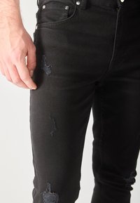 Detailný záber na ruku osoby položenú blízko stehna, oblečenú v čiernych poškodených džínsoch s malými roztrhnutiami.