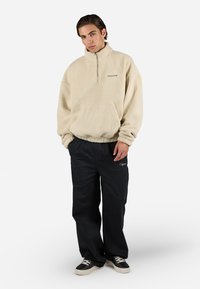 Beige fleece pullover with half-zip and front pocket; paired with black trousers featuring a subtle logo and black sneakers. Smooth texture.