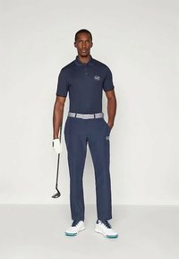 Ναυτικό πόλο μπλουζάκι με λογότυπο EA7, σε συνδυασμό με ταιριαστά παντελόνια. Το σύνολο διαθέτει μία γκρι ζώνη και λευκά παπούτσια γκολφ με μπλε λεπτομέρειες.