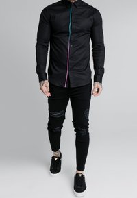 Schwarzes Hemd mit Knopfleiste, das eine türkis- und pinkfarbene Kontrastnaht aufweist, kombiniert mit schwarzen, zerrissenen Skinny-Jeans und schwarzen Sneakers.