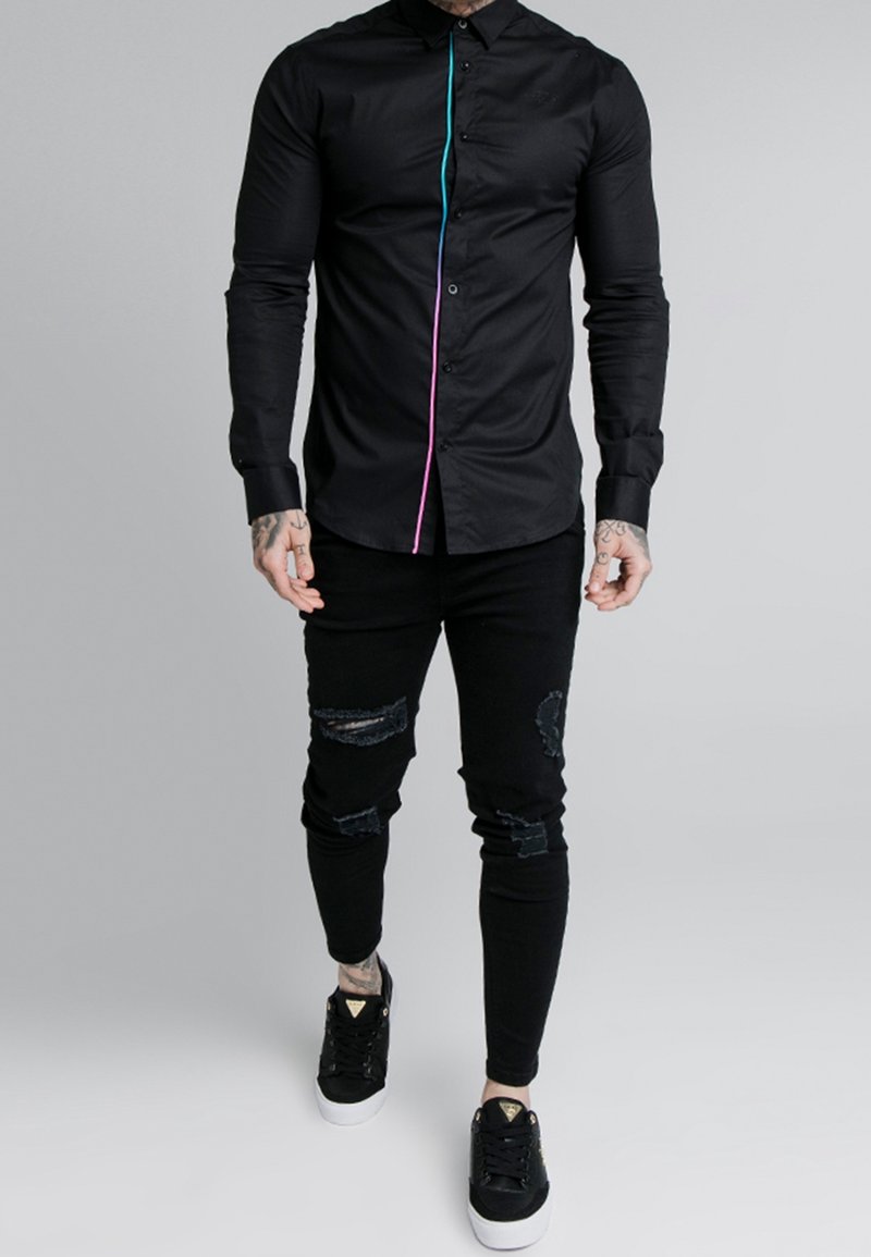 Schwarzes Hemd mit Knopfleiste, das eine türkis- und pinkfarbene Kontrastnaht aufweist, kombiniert mit schwarzen, zerrissenen Skinny-Jeans und schwarzen Sneakers.