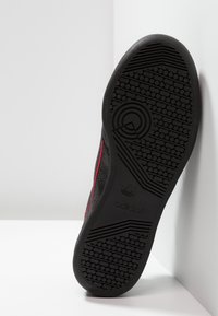 Sort sneakers med en struktureret gummisål, der har røde detaljer langs siden og et præget logo på sålen.
