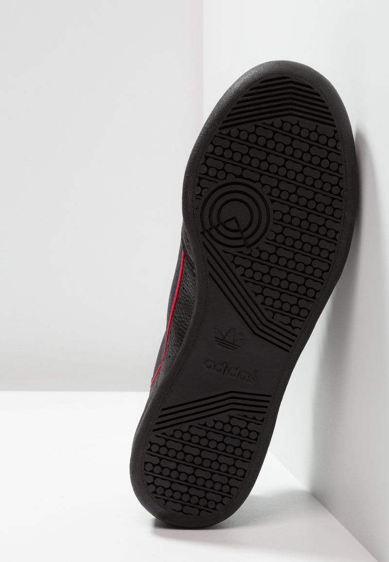 Zapatilla negra con una suela de goma texturizada, con acentos rojos a lo largo del lateral y un logo en relieve en la suela.