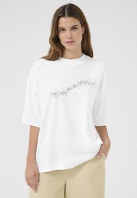 Witte oversized t-shirt met korte mouwen en een subtiel zwart gespikkeld patroon op de borst. Zachte katoen materiaal, relaxte pasvorm.