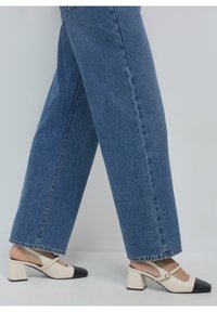 Des escarpins à talon slingback bicolores noir et blanc avec un bout carré, un talon carré, et une lanière avec une boucle en or, associés à un jean large bleu.