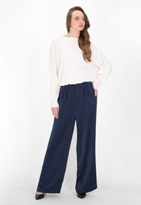 Zwarte baggy broek met plooien en een losse witte langemouwen top. Zwarte hoge hakken maken de look compleet.