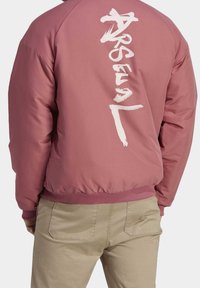 Ljusrosa bomberjacka med ribbade muddar och nederkant, med stor vit text på baksidan. Slätt syntetiskt material med en avslappnad passform.