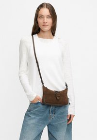Borsa a tracolla in suede marrone con forma piatta, tracolla regolabile e dettaglio in metallo. Indossata con una camicia bianca a maniche lunghe e jeans blu.