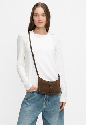 Braune Wildleder-Umhängetasche in flacher Form, mit verstellbarem Riemen und einem Metallschnallendetail. Getragen mit einem weißen Langarmshirt und blauen Jeans.