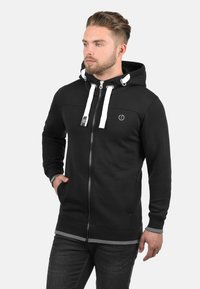Schwarze Zip-Hoodie mit weißen Kordeln und Logo, mit einem einfarbigen Design, Kängurutaschen und einem gerippten Saum, getragen über dunklen Jeans.