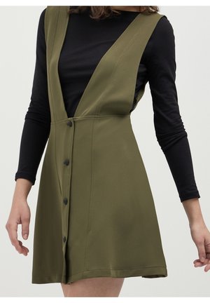 Kvinde iført ærmeløs olivengrøn kjole med knapper foran over en sort top med lange ærmer, stående foran en ensfarvet lys baggrund.