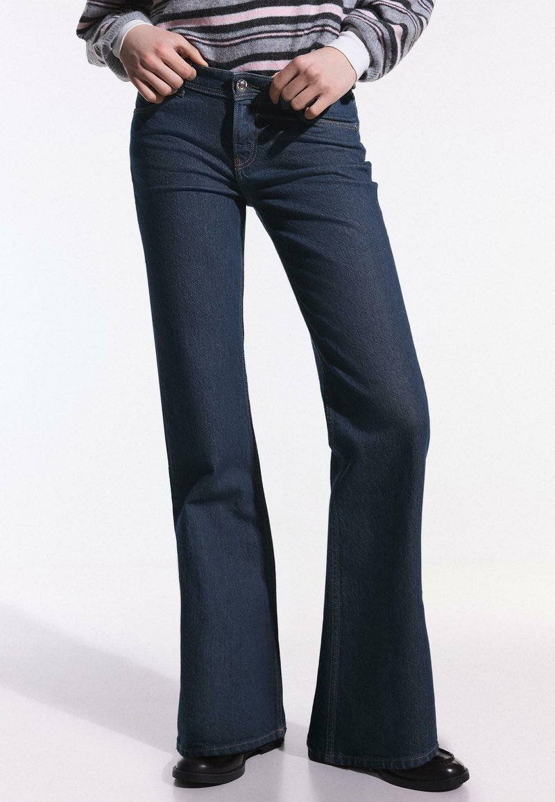 Personne portant un jean évasé bleu foncé et des chaussures noires, ajustant la taille d'un pull rayé rentré dans le jean.