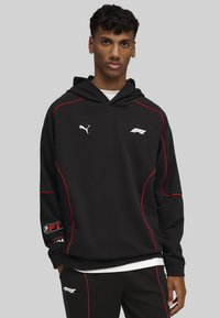 Sort hættetrøje med røde detaljer, der har et Puma-logo og F1-branding. Lange ærmer, snøre i hætten og ribbede manchetter supplerer designet.