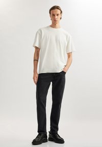 Camiseta de algodón blanca con corte relajado, combinada con pantalones de mezclilla negros y zapatos de cuero negro, de pie frente a un fondo claro.