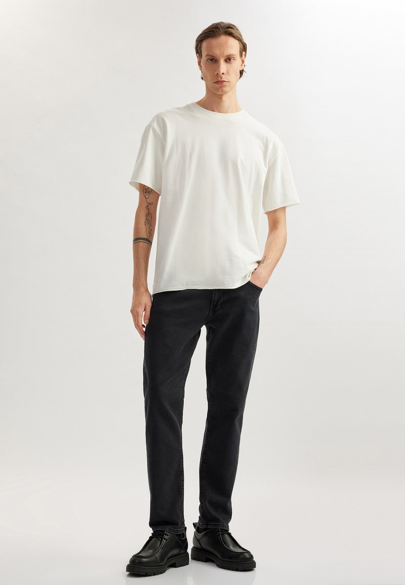 Camiseta de algodón blanca con corte relajado, combinada con pantalones de mezclilla negros y zapatos de cuero negro, de pie frente a un fondo claro.