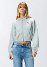 Felpa corta grigio chiaro con cerniera frontale, dotata di una tasca frontale e una toppa a forma di cuore rosso. Abbinata a jeans blu a vita alta.