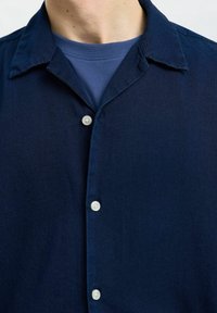 Mörkblå skjorta med knappar och spridningskrage, med vita knappar och en mjuk tygstruktur. Bärs ovanpå en blå t-shirt med rund hals.