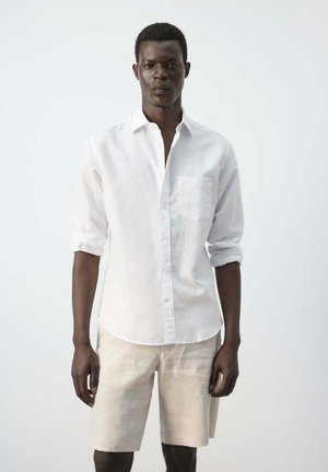 Hombre de piel oscura con camisa blanca de manga larga abotonada y pantalones cortos beige claro, de pie frente a un fondo liso y claro.