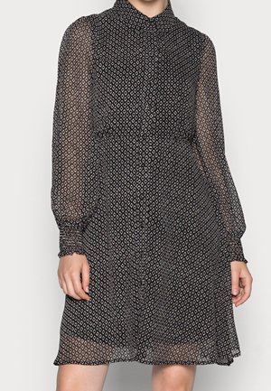 Schwarz-weiß gemustertes langärmeliges Kleid mit Knopfleiste vorn, transparenten Ärmeln und tailliertem Schnitt, getragen von einer stehenden Person.