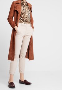 Brun ruskinds trenchcoat, leopardmønstret bluse, beige tætsiddende bukser og burgunderrøde loafers. Glatte teksturer og elegante designelementer.